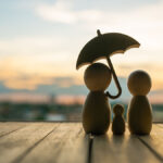 backside-of-three-simple-wooden-figures-representing-a-family-with-an-umbrella-facing-towards-a-beach-sunset