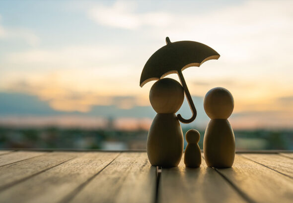backside-of-three-simple-wooden-figures-representing-a-family-with-an-umbrella-facing-towards-a-beach-sunset